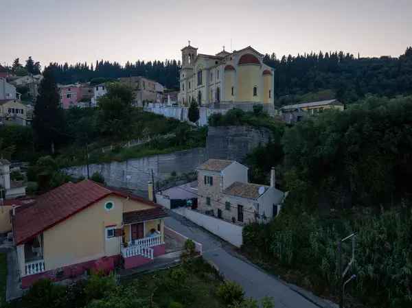 Huzurlu bir köy sahnesinde tepede yükselen bir kilise var. Geleneksel evler ve yol yemyeşil ağaçların arasına yerleşmiş..