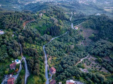 Yüksek açılı bir çekimde yoğun bitki örtüsünün içinde bina ve evlerle çevrili bir yol görülüyor. Manzara canlı, doğal bir görüntü sergiliyor..