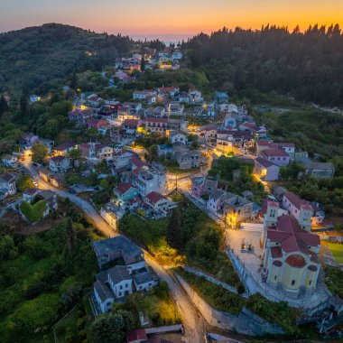 Alacakaranlıkta yemyeşil bir yamaçta, okyanus kıyısında turuncu ışıklı büyüleyici bir köy manzarası.. 