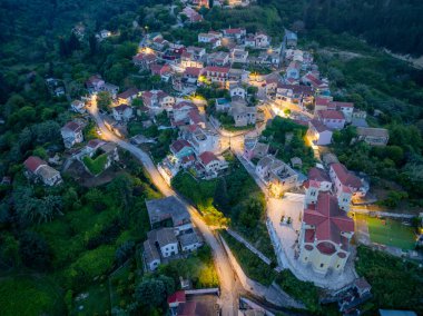 Yukarıdan çekilen sakin bir köy sahnesinde, yemyeşil evlerin arasına yerleşmiş, akşam ışıklarının yumuşak parıltısıyla yıkanmış evler gözler önüne seriliyor..