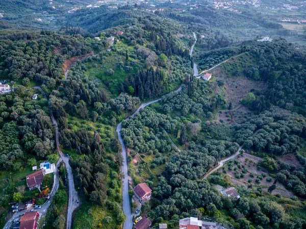 Yüksek açılı bir çekimde yoğun bitki örtüsünün içinde bina ve evlerle çevrili bir yol görülüyor. Manzara canlı, doğal bir görüntü sergiliyor..
