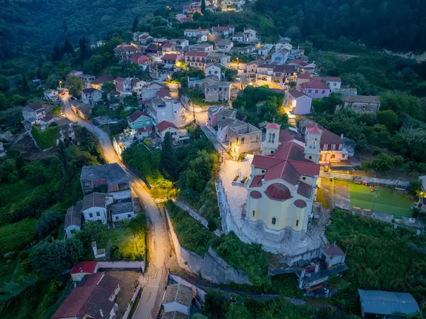 Bir köyün gösterişli evlerinin yüksek açılı görüntüsü ve kırmızı kiremit çatılı bir kilise, ağaçlarla vurgulanmış ve alacakaranlıkta sokak lambalarıyla aydınlatılmış..
