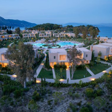 Günbatımında lüks bir tatil köyünü gösteren yüksek açılı bir manzara, modern binalar, manzaralı bahçeler ve kıyı şeridi yakınında büyük bir havuz..