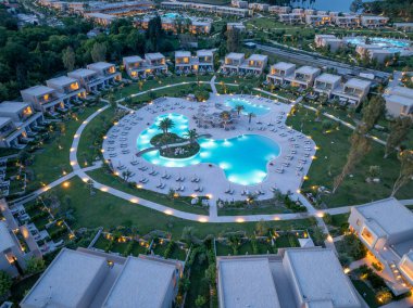 Havadan çekilen bir görüntü, alacakaranlıkta lüks bir tatil köyünü, merkezi havuzu ve manikürlü manzarayı çevreleyen villaları gösteriyor. Sahne sükunet yayar..