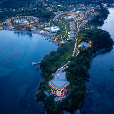 Bir hava görüntüsü, akşam karanlığında aydınlanan ve bereketli manzaralar ve sakin sularla bezenmiş pitoresk bir sahil şeridi boyunca uzanan bir sahil beldesi yakalar..