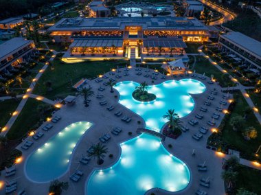 Bu resim alacakaranlıktaki lüks bir otel kompleksini çekiyor, büyüleyici mimarisi, aydınlık havuzları, güneşli yatakları ve canlı bitki örtüsüyle..