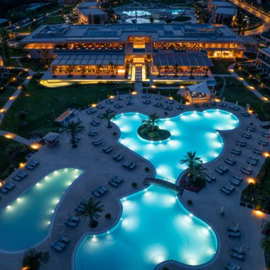 Gece görüşünde birbirine bağlı havuzlar, palmiye ağaçları ve tepesinde açık hava yemekleri olan büyük bir bina sergileniyor..