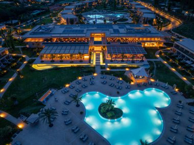 Bu hava görüntüsü, büyük havuzunu, palmiye ağaçlarını, modern mimariyi ve hareketli ambiyansını vurgulayarak gece güzel aydınlatılmış bir tatil köyünü yakalar..