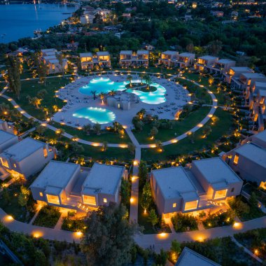 Yüksek açılı çekimler bir otel tatil beldesinin alacakaranlıkta aydınlatıldığı, binalar ve yeşil manzaralarla çevrili çok sayıda havuzların yer aldığı bir sergi..