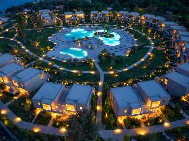 Bir hava çekimi alacakaranlıktaki bir tatil köyünü gösteriyor. Modern villalar, dikkatlice tasarlanmış peyzaj ortasındaki aydınlık havuzların etrafını sarar..