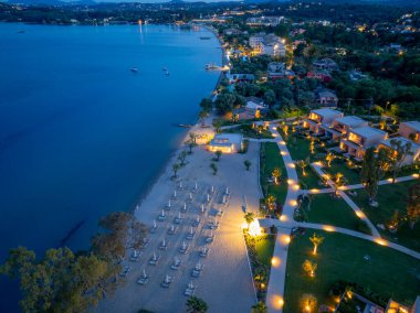 Hava manzarası, alacakaranlıkta huzurlu bir sahil kenarı tatil beldesi, aydınlık yollar, modern villalar ve sakin sahil manzarası gösteriyor..