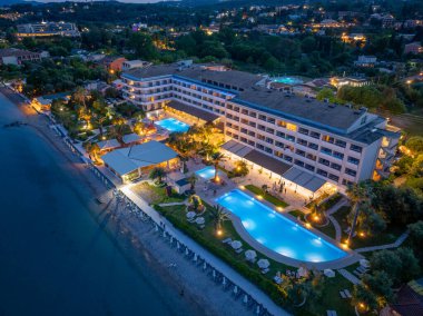 Havuz, palmiye ağaçları ve yakındaki sahil şeridini aydınlatan ışıklarla dolu çok katlı bir sahil otelinin gece görüntüsü..