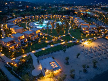 Havuz etrafında, aydınlık yolları ve plaja erişimi olan dairesel bir villa düzeninin yer aldığı lüks bir tatil köyünün gece hava görüntüsü..