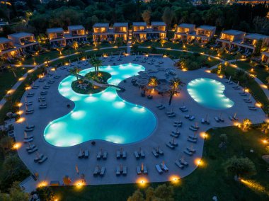 Yüksek bir manzara, gece güneşli yatakları ve dolambaçlı havuzu olan bir tatil köyünü gösteriyor. Villalar, palmiye ağaçları ve zemin ışıkları manzarayı kaplıyor..