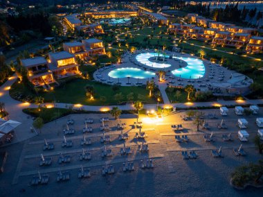 Bu hava görüntüsü alacakaranlıkta gösterişli bir tatil köyü yakalıyor. Aydınlatılmış havuzları, zarif villaları ve güneşli yataklı huzurlu plaj alanını aydınlatıyor..