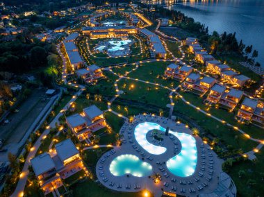 Parlak ışıklı havuzlar, özel villalar ve titizlikle döşenmiş bahçelerle dolu lüks bir tatil köyünü gece gösteren hava görüntüsü..