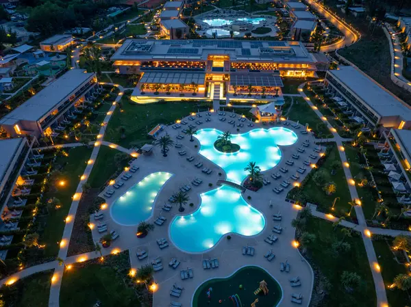 Gece çekimleri, aydınlık havuzları, modern mimarisi, palmiye ağaçları ve manikürlü manzarası olan bir tatil köyünü gösteriyor. Huzurlu, lüks tatil ortamı.