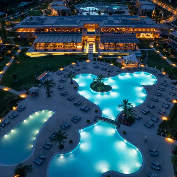 Gece görüşünde birbirine bağlı havuzlar, palmiye ağaçları ve tepesinde açık hava yemekleri olan büyük bir bina sergileniyor..