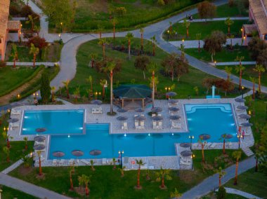 Palmiye ağaçları ve kaldırımlarla çevrili yüzme havuzlarını gösteren yüksek açılı bir çekim. Bir çardak, yemyeşil bir tatil köyünde iki havuz arasında duruyor..