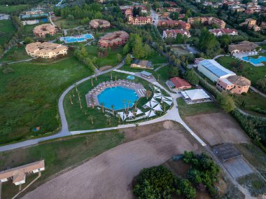 Daire şeklinde bir havuz, palmiye ağaçları ve çeşitli konforların yer aldığı, binalar ve yeşillikler içinde geçen sakin bir tatil beldesinin havadan çekilmiş görüntüsü..