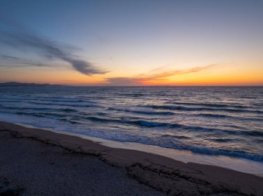 Gün batımında sakin bir okyanusu gösteren sakin bir sahil manzarası, yumuşak dalgaların canlı, renkli bir gökyüzünün altında kumlu sahile vurduğu.