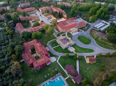 Ağaçlarla çevrili, klasik kırmızı kiremitli çatıları olan bir tatil köyünün çarpıcı bir görüntüsü. Havuz manzarayı güzelleştirir..