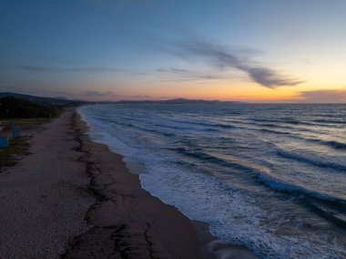 Bir hava manzarası, alacakaranlıkta sakin bir sahili yakalar, yumuşak dalgaların kıyıya vurduğu, güneşin sıcacık parıltısıyla yıkandığı,.