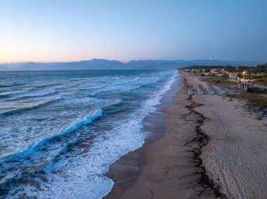 Bu hava görüntüsü, sakin bir kıyı şeridini, yumuşak dalgaların kumlu bir kumsalla, uzak dağlarla buluşmasını ve alacakaranlıkta huzurlu bir atmosferi yakalar..