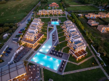 Havadan bakıldığında, simetrik mimari, ışıltılı havuzlar ve düzgün desenli peyzaj tasarımı içeren organize lüks bir tatil köyü sergileniyor..