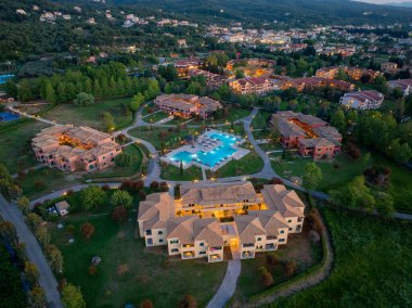 Parlayan binalar, parlak bir havuz ve yemyeşil ve uzak bir kasabaya karşı kurulmuş dolambaçlı yollar ile aydınlık bir tatil köyünü görüntüler..