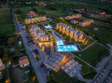 Birden fazla villası olan bir tatil köyünün yüksek açılı görüntüsü, bağlı havuzlar, bir restoran ve plaj girişi, akşam gökyüzünün altında aydınlatılmış..