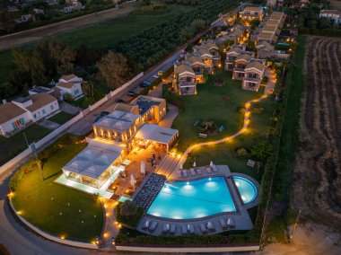 Havadan çekilen bir fotoğraf birçok villa, bir havuz ve parlayan yürüyüş yollarının olduğu bir tatil köyü sunar, alacakaranlık çöktüğünde sakin atmosferi yakalar..