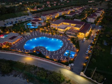 Gece boyunca mimarisini, havuzlarını ve manzarasını sergileyen bir tatil köyünün yüksek açılı görüntüsü. Binalar ve park halindeki arabalar aydınlanıyor..