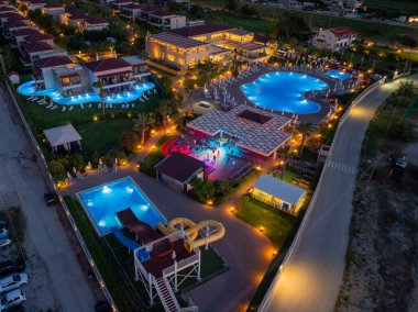 Hava manzarası, aydınlık havuzlar, su kaydırakları ve hareketli bir açık hava toplanma alanının yer aldığı canlı bir tatil beldesi gösteriyor..