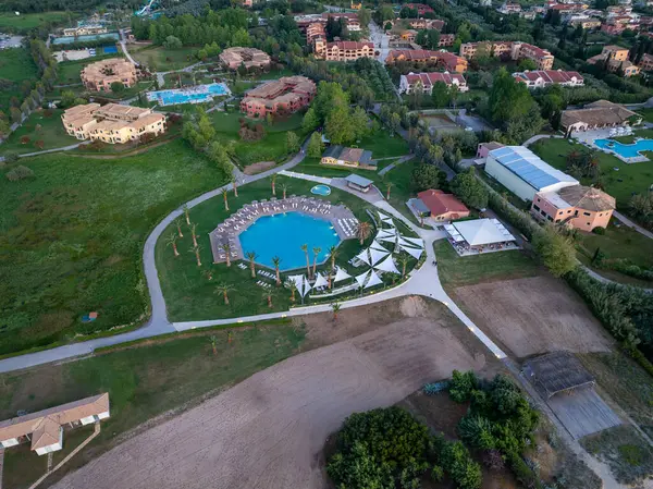 Daire şeklinde bir havuz, palmiye ağaçları ve çeşitli konforların yer aldığı, binalar ve yeşillikler içinde geçen sakin bir tatil beldesinin havadan çekilmiş görüntüsü..