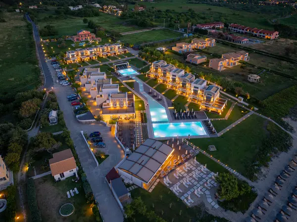 Birden fazla villası olan bir tatil köyünün yüksek açılı görüntüsü, bağlı havuzlar, bir restoran ve plaj girişi, akşam gökyüzünün altında aydınlatılmış..