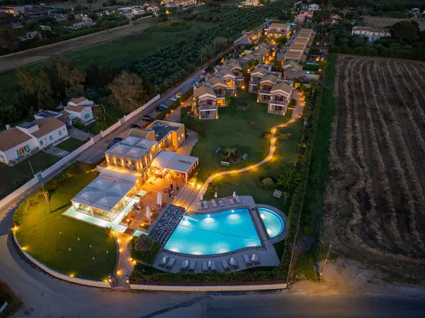 Bir hava görüntüsü alacakaranlıkta aydınlık villaları, bir yüzme havuzu ve yemyeşil ve dingin atmosferde dolambaçlı yolları olan bir tatil beldesi sergiliyor..