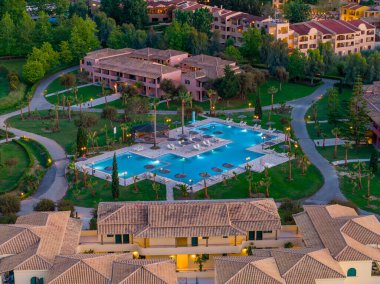 Büyük, mavi bir yüzme havuzu, güneş pansiyonları, yürüyüş yolları ve çevreleyen yeşil çimenler ve ağaçların ortasındaki binaların yer aldığı bir tatil köyünü gösteren bir hava görüntüsü..