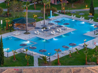 Yükseltilmiş bir perspektif, güneş yatakları, şemsiyeler, yemyeşil bitki örtüsü ve gölgeli bir kulübe yapısı ile birlikte genişleyen bir yüzme havuzu kompleksi yakalar..