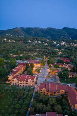 Terracotta çatıları olan, yeşillik içinde yuvalanmış, akşam karanlığında hafif bir parıltıyla aydınlanan bir tatil köyünü bir hava manzarası yakalar..