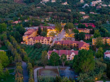 Havadan çekilen bir görüntü, kendine özgü kırmızı kiremitli çatıları olan, sakin ve davetkar manzaralı yeşil ağaçlarla çevrili bir tatil köyünü yakalar..