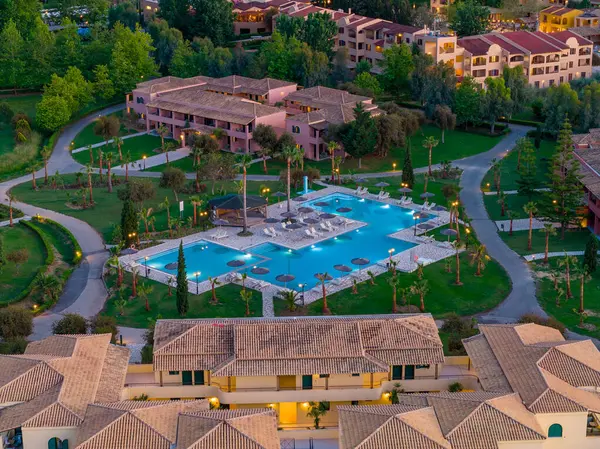 Büyük, mavi bir yüzme havuzu, güneş pansiyonları, yürüyüş yolları ve çevreleyen yeşil çimenler ve ağaçların ortasındaki binaların yer aldığı bir tatil köyünü gösteren bir hava görüntüsü..