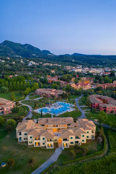 Bu hava fotoğrafında villaları, yüzme havuzu ve palmiyeleri olan bir tatil köyü yer alıyor. Tatil köyü ağaçlardan ve tepelerden oluşan bir arazide..