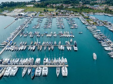 Çeşitli boyutlarda teknelerle dolu büyük bir marina yeşil bir sahil şeridine yakın turkuaz sularda demirledi. Birçok yelkenli kayıkta..