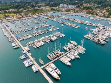 Bir hava sahnesinde çeşitli yelkenliler, katamaranlar ve yatlarla dolu canlı bir marina sahile vuruyor..