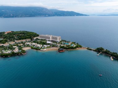 Bir hava görüntüsü, mavi okyanuslar ve uzak dağların yanındaki çeşitli bina ve konforlara sahip bir adada geniş bir tatil köyü kompleksi yakalar..