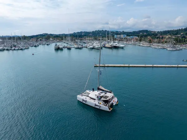Bu görüntüde marinada yüzen bir katamaran var. Birçok gemi demirli. Su mavi ve gökyüzü bulutlu..