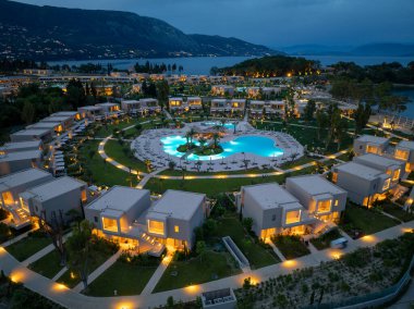 Alacakaranlıkta aydınlık lüks bir tatil köyünün yüksek açılı çekimi, dağlara karşı villaları, havuzu ve manzaralı sahili gösteriyor..