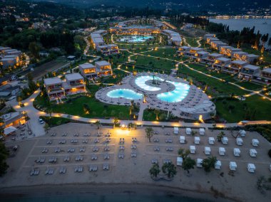 Hava manzarasında modern binalar, büyük bir yüzme havuzu ve geceleri ışıldayan şezlongları olan kumlu bir plaj sergileniyor..