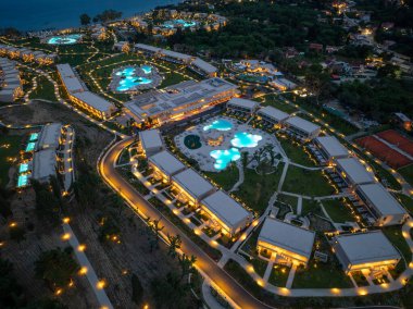 Aydınlatılmış havuzları, modern binaları ve titizlikle döşenmiş arazileri olan geniş bir tatil köyünün gece görüntüsü. Sakin ve lüks bir atmosfer..